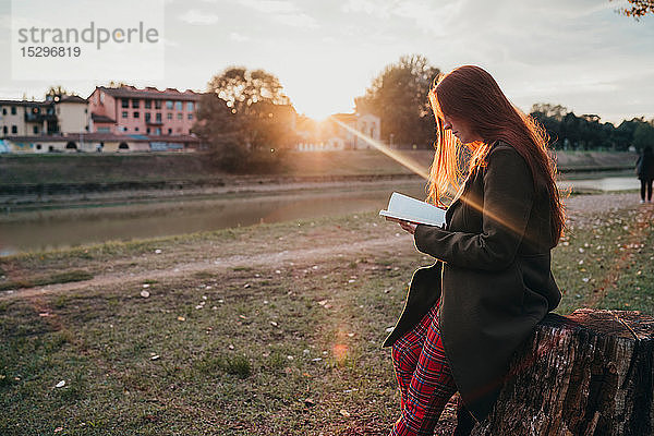 Junge Frau mit langen roten Haaren sitzt auf Baumstumpf und liest am Flussufer bei Sonnenuntergang ein Buch  Florenz  Toskana  Italien