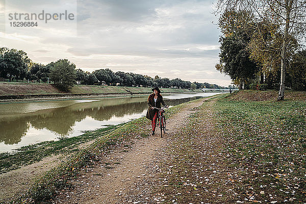 Junge Frau mit langen roten Haaren schiebt Fahrrad am Flussufer  in voller Länge  Florenz  Toskana  Italien