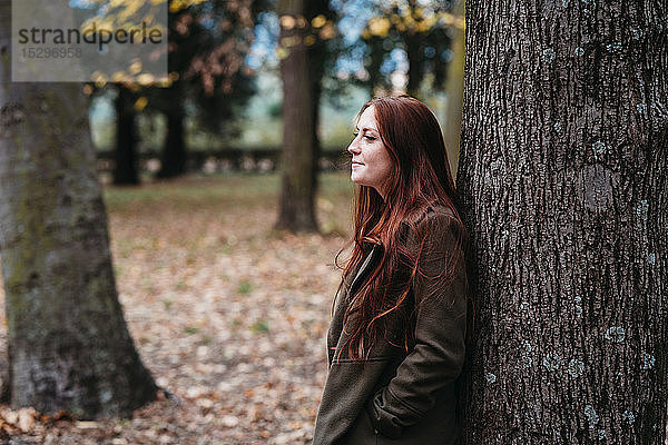 Junge Frau mit langen roten Haaren lehnt an Baumstamm im Herbstpark