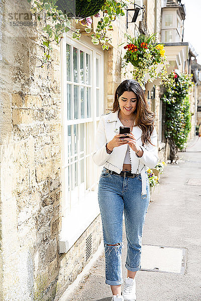 Junge Frau schlendert die Dorfstraße entlang und sieht sich ein Smartphone an  Cotswolds  England