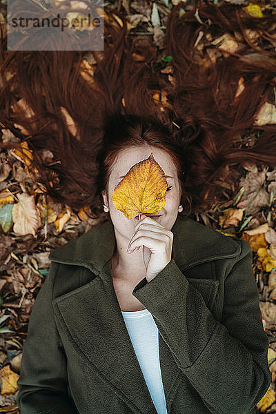 Junge Frau mit langen roten Haaren zwischen Herbstblättern liegend und das Gesicht mit Herbstblatt bedeckend  Überkopfporträt