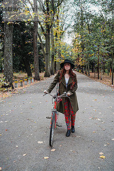 Junge Frau mit langen roten Haaren schiebt Fahrrad im von Bäumen gesäumten Herbstpark  Porträt  Florenz  Toskana  Italien