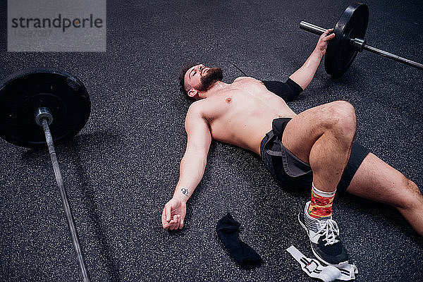 Nacktbrust trainierender junger Mann  der erschöpft zwischen Hanteln in der Turnhalle liegt