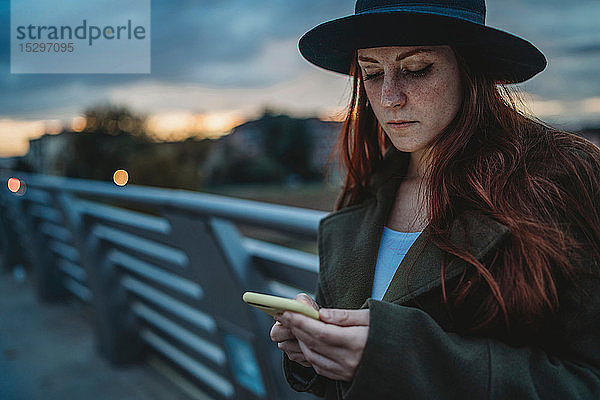 Junge Frau mit langen roten Haaren auf Steg mit Blick auf Smartphone in der Dämmerung  Florenz  Toskana  Italien