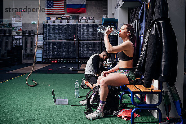 Junge Frau trainiert  sitzt auf einer Bank und trinkt Wasser in Flaschen im Fitnessstudio