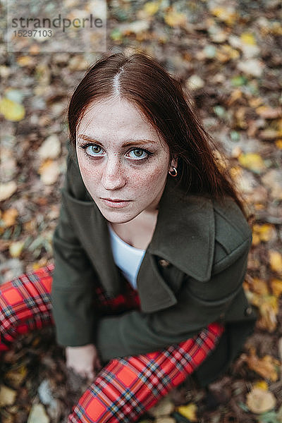Junge Frau mit langen roten Haaren auf Herbstlaub im Park sitzend  Hochwinkelporträt