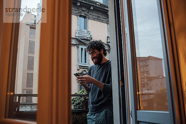 Bärtiger junger Mann benutzt Smartphone auf dem Balkon