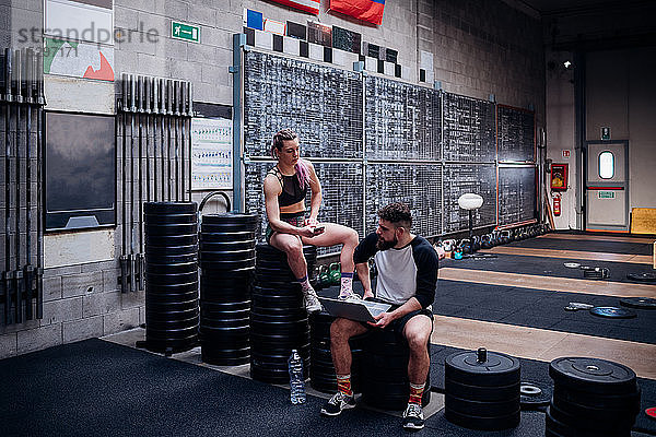 Junge Frau und Mann trainieren gemeinsam im Fitnessstudio  auf Gewichten sitzend mit Smartphone und Laptop