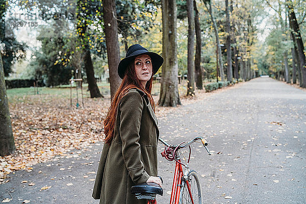 Junge Frau mit langen roten Haaren  die beim Fahrradfahren im Herbstpark über ihre Schulter schaut  Florenz  Toskana  Italien