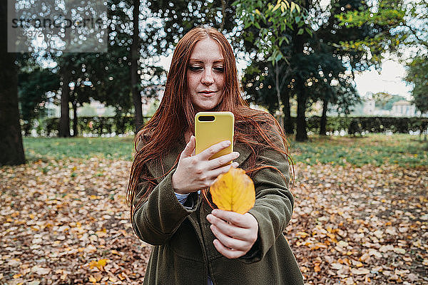 Junge Frau mit langen roten Haaren im Park beim Smartphonefoto ihrer Hand  die ein Herbstblatt hält