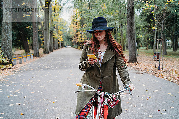 Junge Frau mit langen roten Haaren auf Fahrrad schaut auf Smartphone im Herbstpark  Florenz  Toskana  Italien