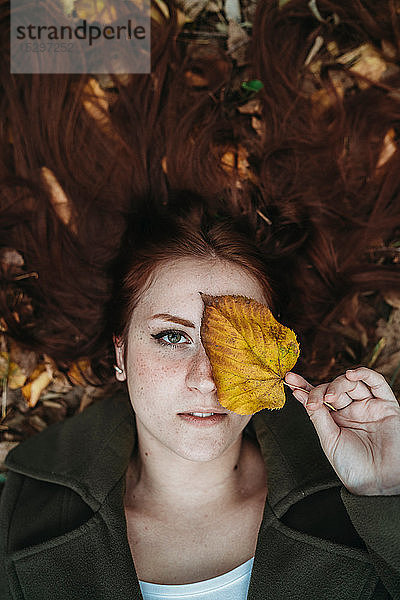 Junge Frau mit langen roten Haaren  zwischen Herbstblättern liegend und das Auge mit Herbstblatt bedeckend  Überkopfporträt