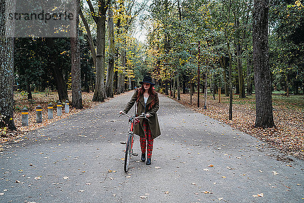 Junge Frau mit langen roten Haaren schiebt Fahrrad im von Bäumen gesäumten Herbstpark  Florenz  Toskana  Italien