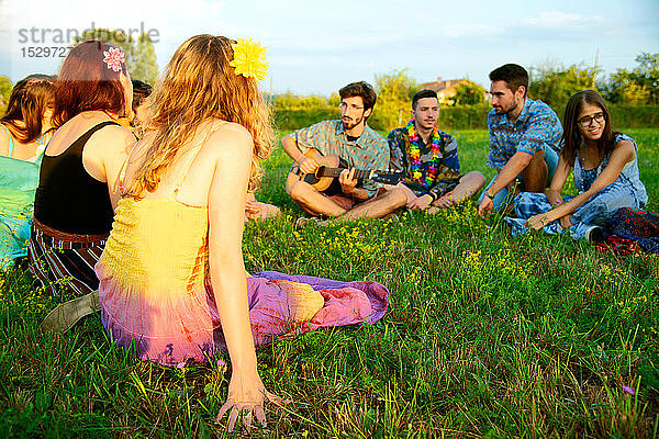 Große Gruppe junger Erwachsener sitzt im Feld und hört akustische Gitarre