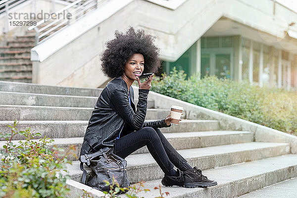 Junge Frau mit Afro-Haaren sitzt auf einer Stadttreppe und spricht mit einem Smartphone