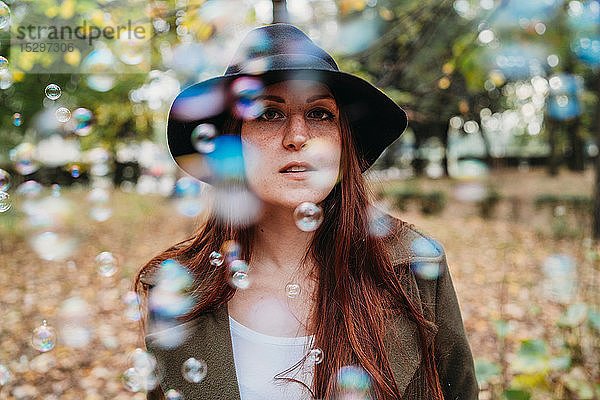 Junge Frau mit langen roten Haaren zwischen schwebenden Blasen im Herbstpark  flach fokussiertes Porträt