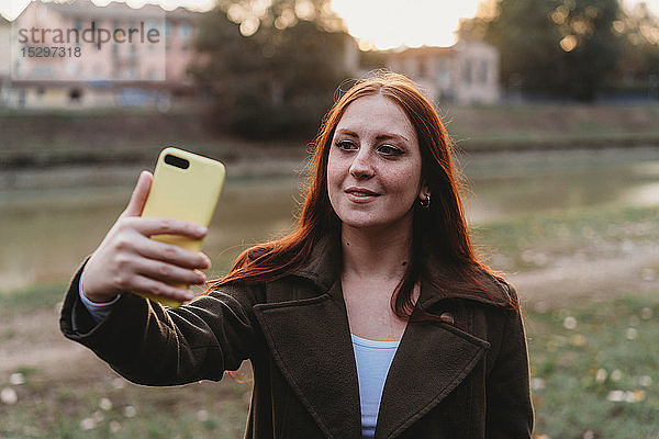 Junge Frau mit langen roten Haaren beim Smartphone-Selfie am Flussufer in der Abenddämmerung  Florenz  Toskana  Italien