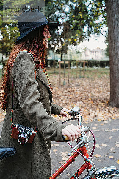 Junge Frau mit langen roten Haaren schiebt Fahrrad im Herbstpark