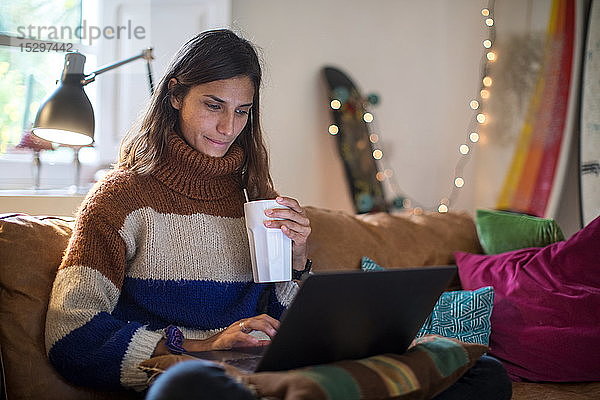 Junge Frau in Strickpulli auf Wohnzimmersofa mit Getränk und Laptop