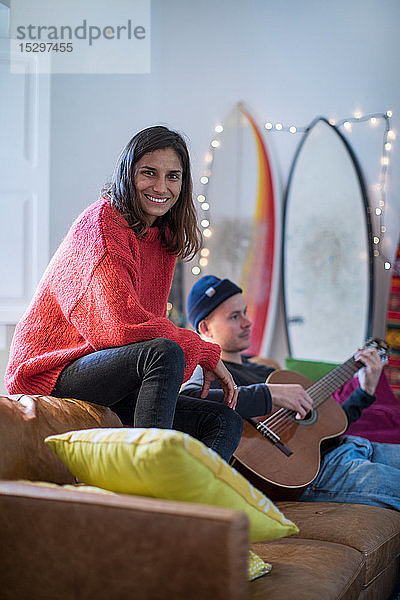 Junge Frau sitzt auf einem Wohnzimmersofa und ein junger Mann spielt Gitarre