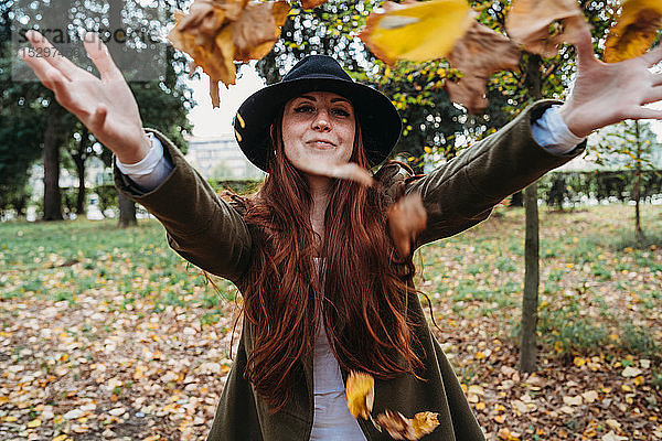 Junge Frau mit langen roten Haaren wirft Herbstblätter im Park