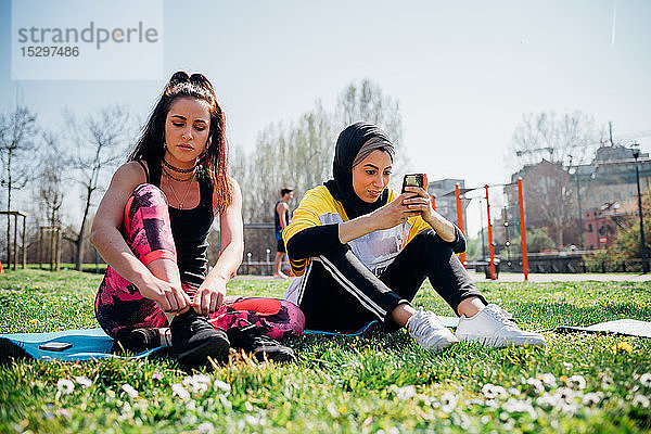 Calisthenics-Kurs im Fitnessstudio im Freien  junge Frauen sitzen auf Gras  binden Schnürsenkel und schauen auf das Smartphone