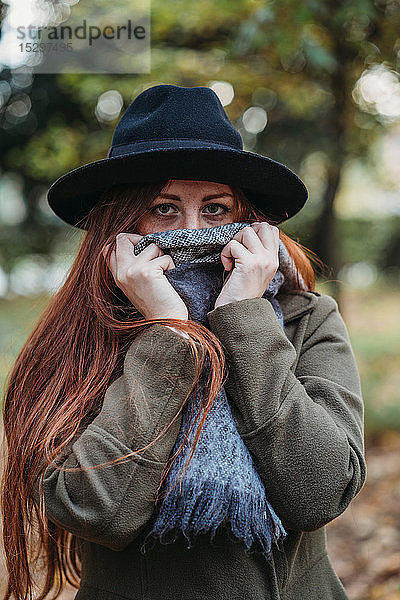 Junge Frau mit langen roten Haaren im Herbstpark  den Mund mit einem Schal bedeckend  Porträt