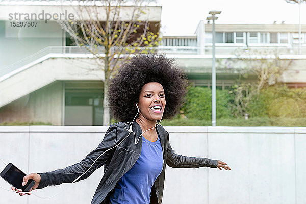 Glückliche junge Frau mit Afrofrisur in der Stadt  die lacht und zu Smartphone-Musik tanzt