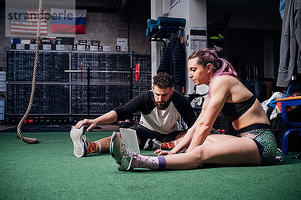 Junge Frau und Mann trainieren gemeinsam im Fitnessstudio  sitzen auf dem Boden  machen Aufwärmübungen und schauen auf den Laptop