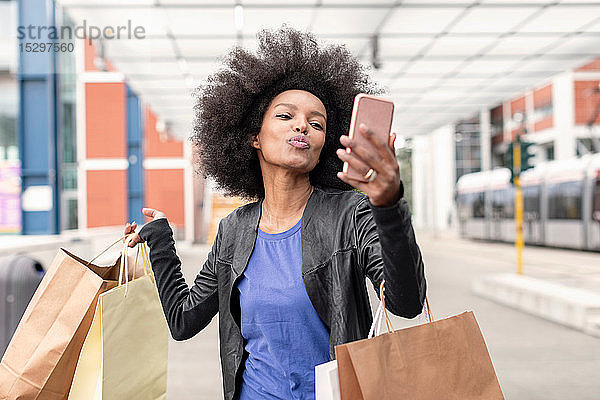 Junge Frau mit Afro-Haaren am Stadtbahnhof  die Einkaufstaschen trägt  mit spitzen Lippen für Smartphone-Selfie