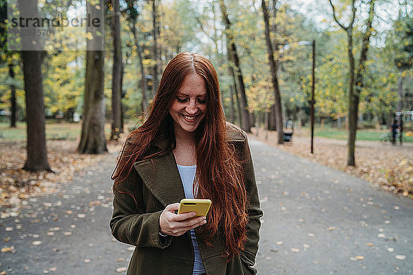 Junge Frau mit langen roten Haaren betrachtet Smartphone im von Bäumen gesäumten Herbstpark  Florenz  Toskana  Italien