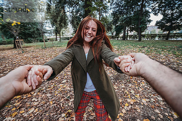 Junge Frau mit langen roten Haaren tanzt  während sie die Hände ihres Freundes im Herbstpark hält  persönliche Perspektive