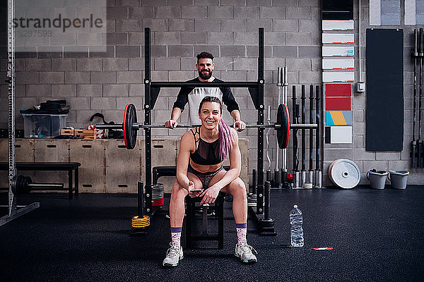 Junge Frau und Mann trainieren gemeinsam im Fitnessstudio  sitzen auf Hantelbank  Porträt