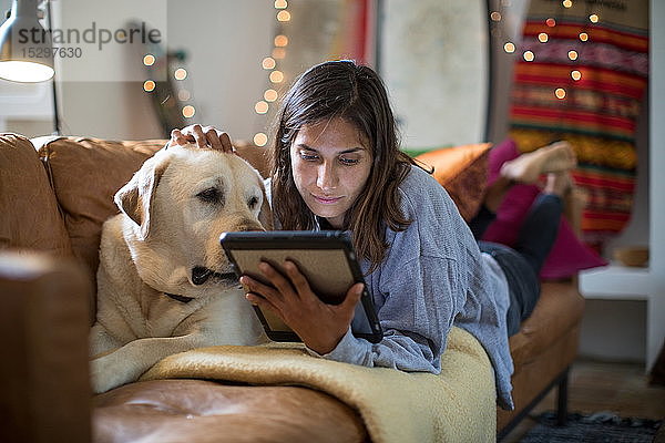 Labrador Retriever und junge Frau liegen auf einem Wohnzimmersofa und schauen auf ein digitales Tablett