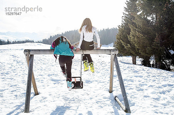 Zwei jugendliche Skifahrerinnen auf der Schaukel in schneebedeckter Landschaft  Rückansicht  Tirol  Steiermark  Österreich