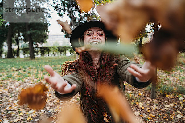 Junge Frau mit langen roten Haaren wirft Herbstblätter im Park