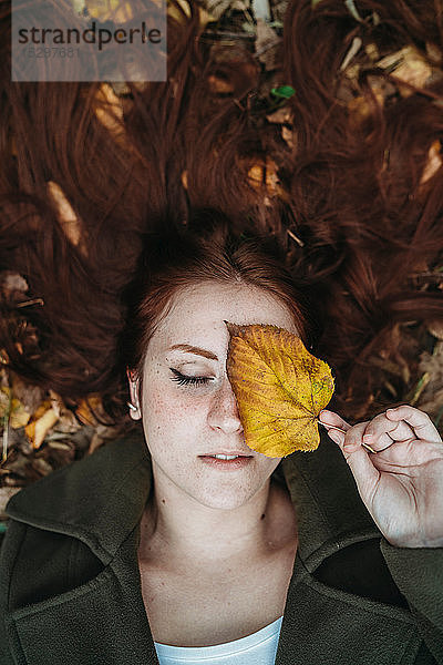 Junge Frau mit langen roten Haaren  zwischen Herbstblättern liegend und das Auge mit Herbstblatt bedeckend  Überkopfporträt