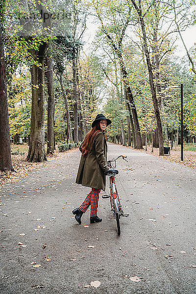 Junge Frau mit langen roten Haaren  die über ihre Schulter schaut  während sie im Herbst im Park ein Fahrrad schiebt  in voller Länge  Florenz  Toskana  Italien