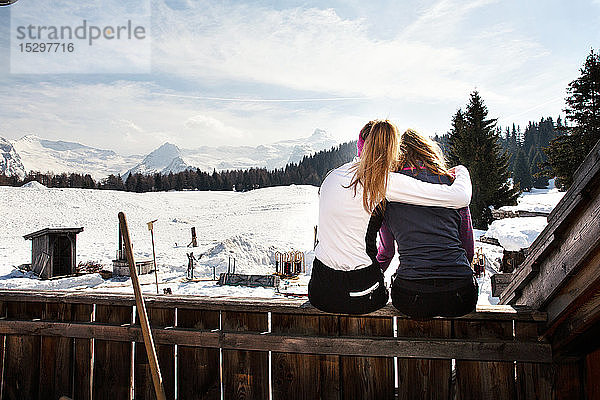 Zwei jugendliche Skifahrerinnen sitzen auf der Dachterrasse einer Hütte in schneebedeckter Landschaft  Rückansicht  Tirol  Steiermark  Österreich