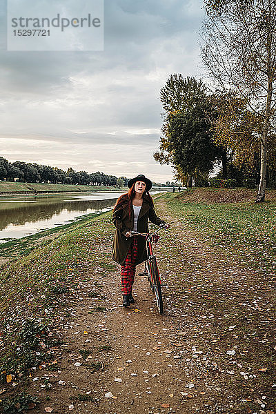 Junge Frau mit langen roten Haaren  die am Flussufer ein Fahrrad schiebt  Porträt in voller Länge  Florenz  Toskana  Italien