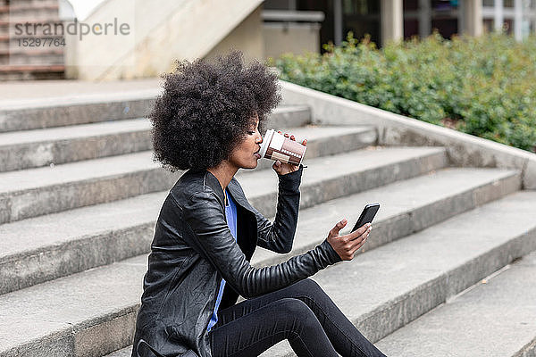 Junge Frau mit Afro-Haaren sitzt auf einer Stadttreppe  trinkt Kaffee zum Mitnehmen und schaut auf ein Smartphone