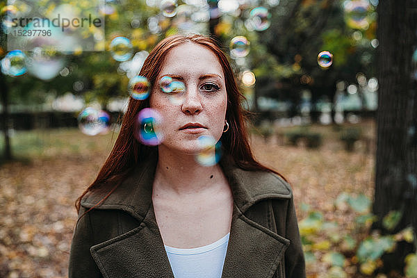 Junge Frau mit langen roten Haaren zwischen schwebenden Blasen im Herbstpark  flach fokussiertes Porträt