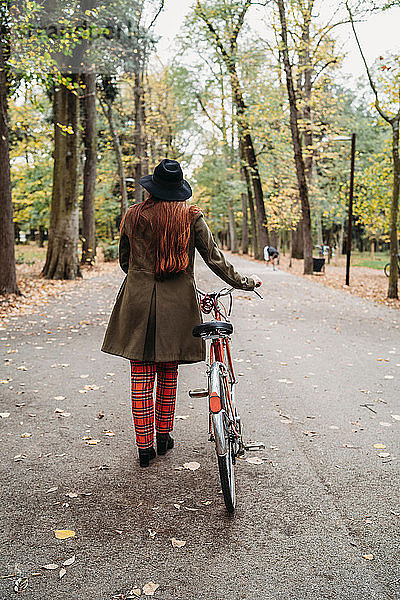 Junge Frau mit langen roten Haaren schiebt Fahrrad im Herbstpark  Rückansicht in voller Länge  Florenz  Toskana  Italien