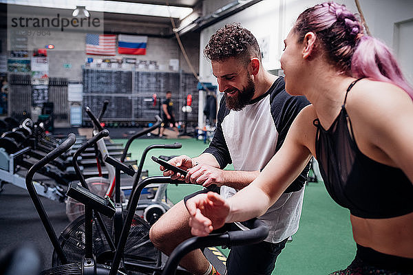 Junge Frau und Mann trainieren gemeinsam auf Fitness-Fahrrädern  Blick auf Smartphone