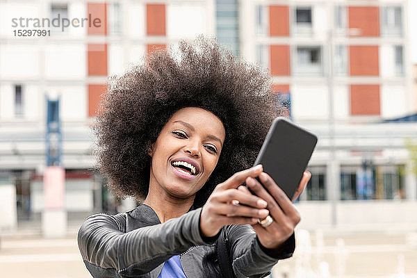 Glückliche junge Frau mit Afro-Haaren in der Stadt  die Smartphone-Selfie