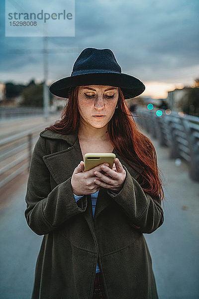 Junge Frau mit langen roten Haaren auf Steg schaut in der Dämmerung auf Smartphone