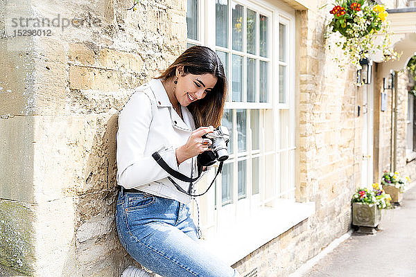 Junge Frau lehnt an Straßenmauer und betrachtet Fotos mit Digitalkamera  Cotswolds  England
