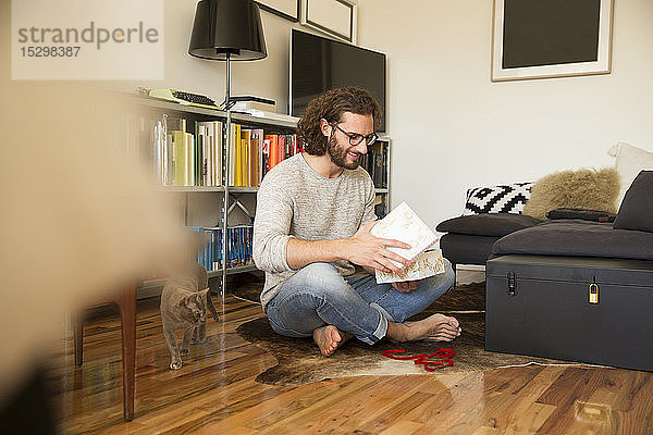 Junger Mann sitzt auf dem Boden im Wohnzimmer und packt ein Geschenk aus