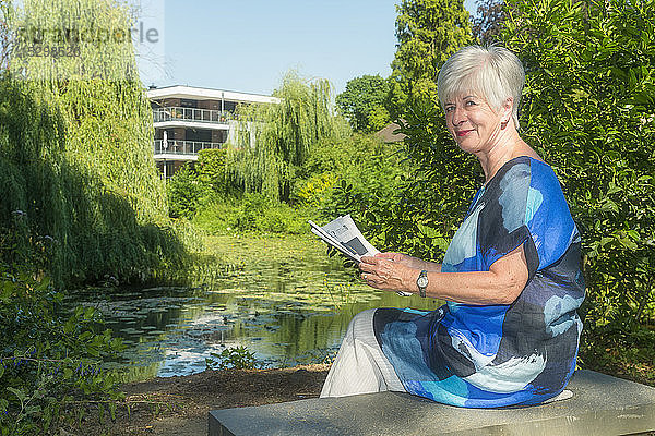 Porträt einer lächelnden älteren Frau  die mit Zeitung am See sitzt