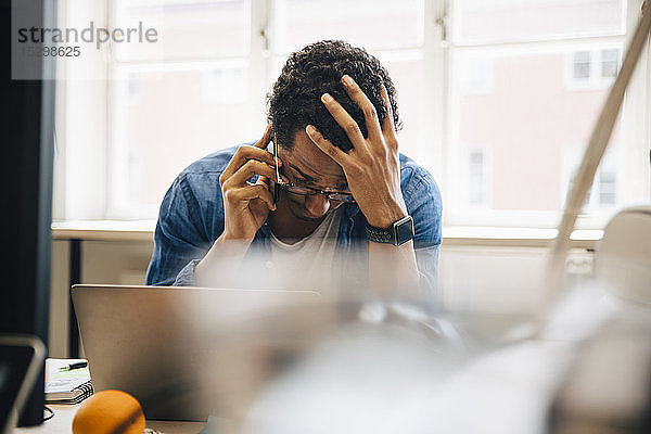 Frustrierter männlicher Computer-Hacker mit der Hand im Haar  der mit einem Smartphone telefoniert  während er den Laptop im Kreativbüro benutzt